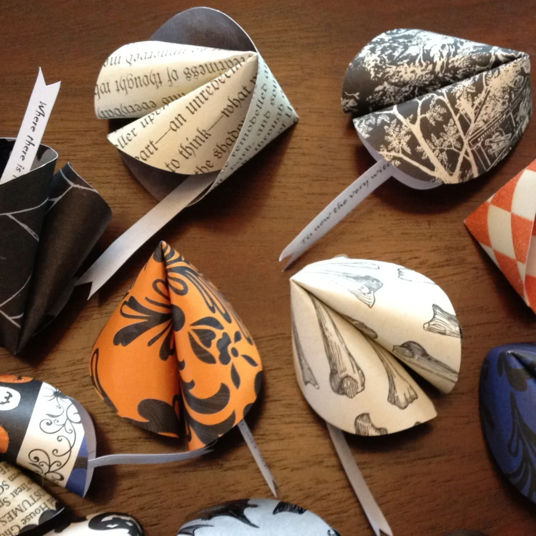 Colorful folded-paper heart ornaments made from book pages and patterned paper are scattered across a wooden table, creating a playful handmade craft display.
