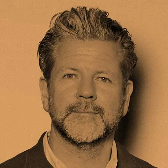 Sepia-toned portrait of a middle-aged man with tousled hair and a short beard, wearing a dark jacket; he looks calmly at the camera.