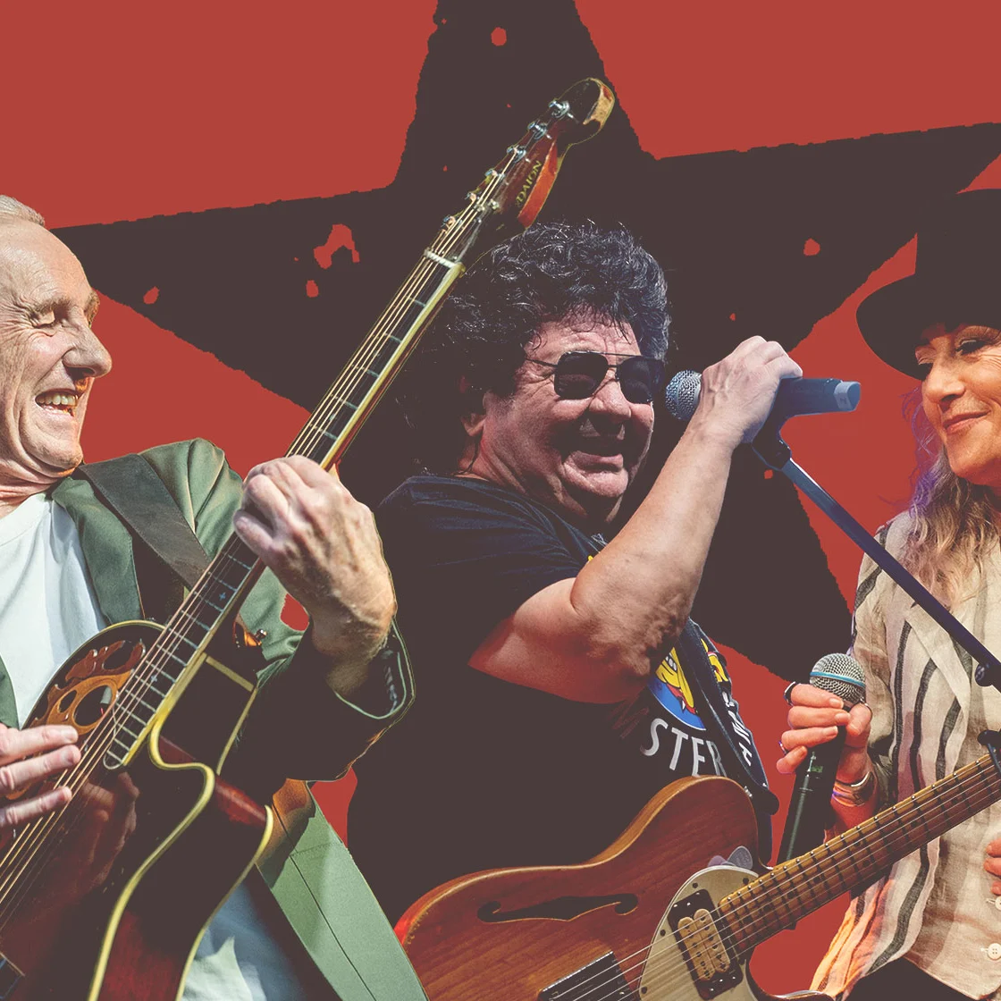 Three musicians perform against a red backdrop: a gray-haired guitarist, a curly-haired singer at a microphone, and a woman in a black hat with a guitar; lively, celebratory tone.