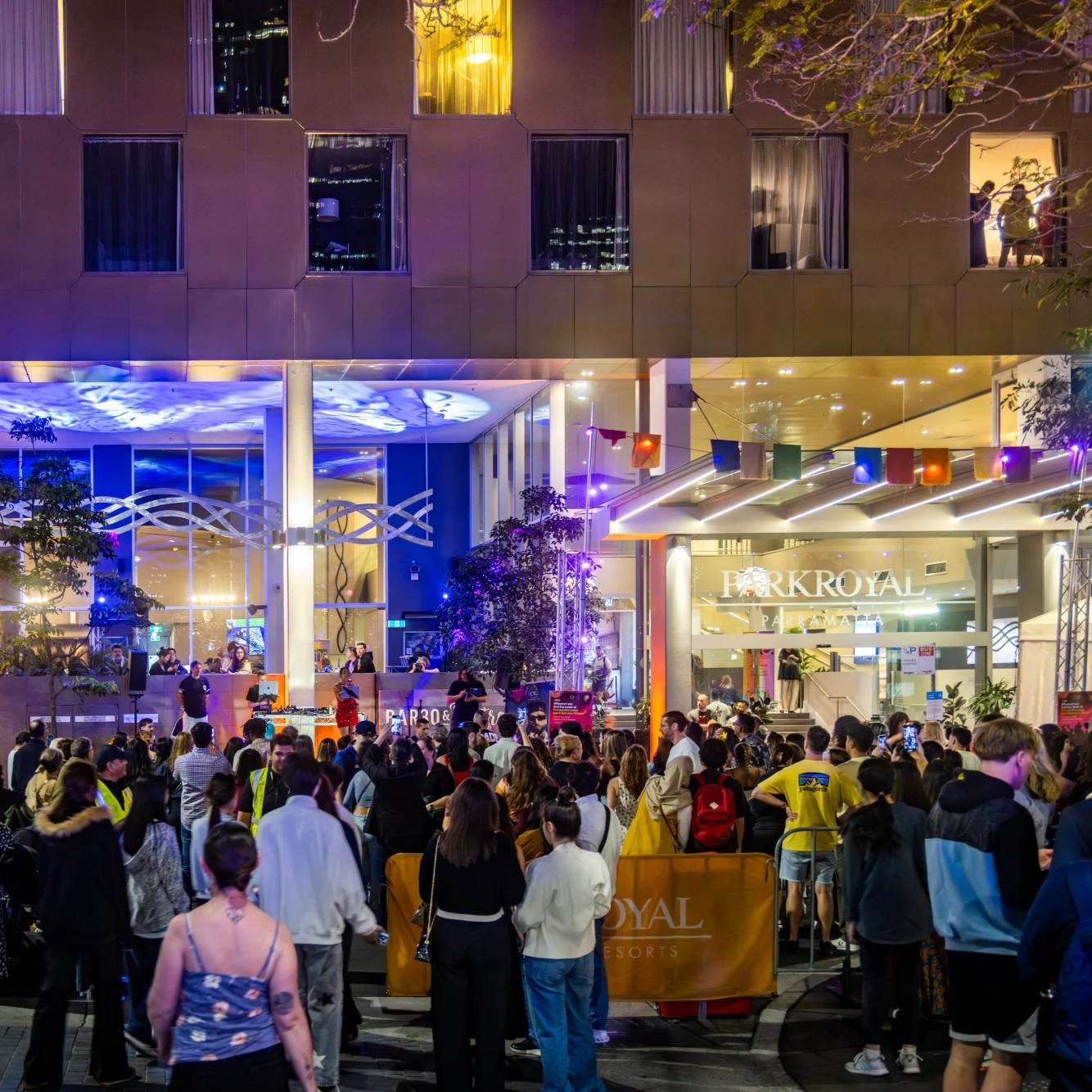 A large evening crowd gathers outside the brightly lit Park Royal entrance, where blue and purple lights illuminate the modern facade, creating a lively event atmosphere.