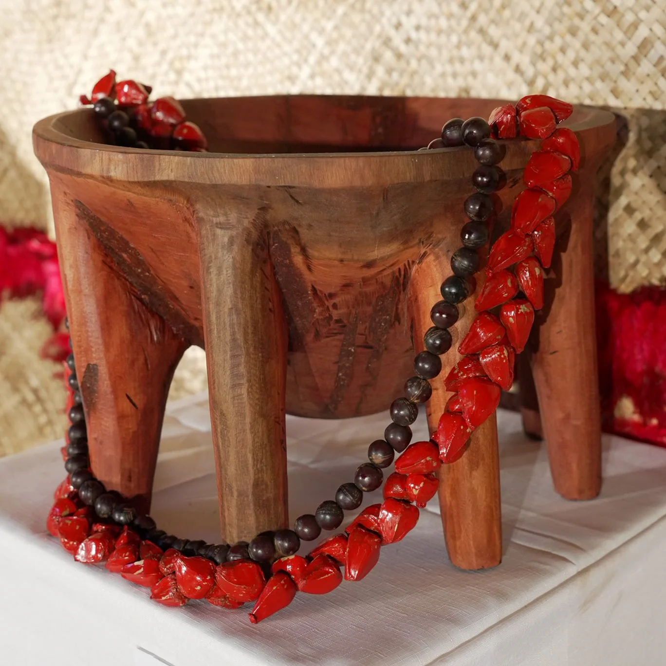 A rustic wooden three-legged bowl stands on a white display, draped with red coral beads and a dark bead necklace against woven fabric.