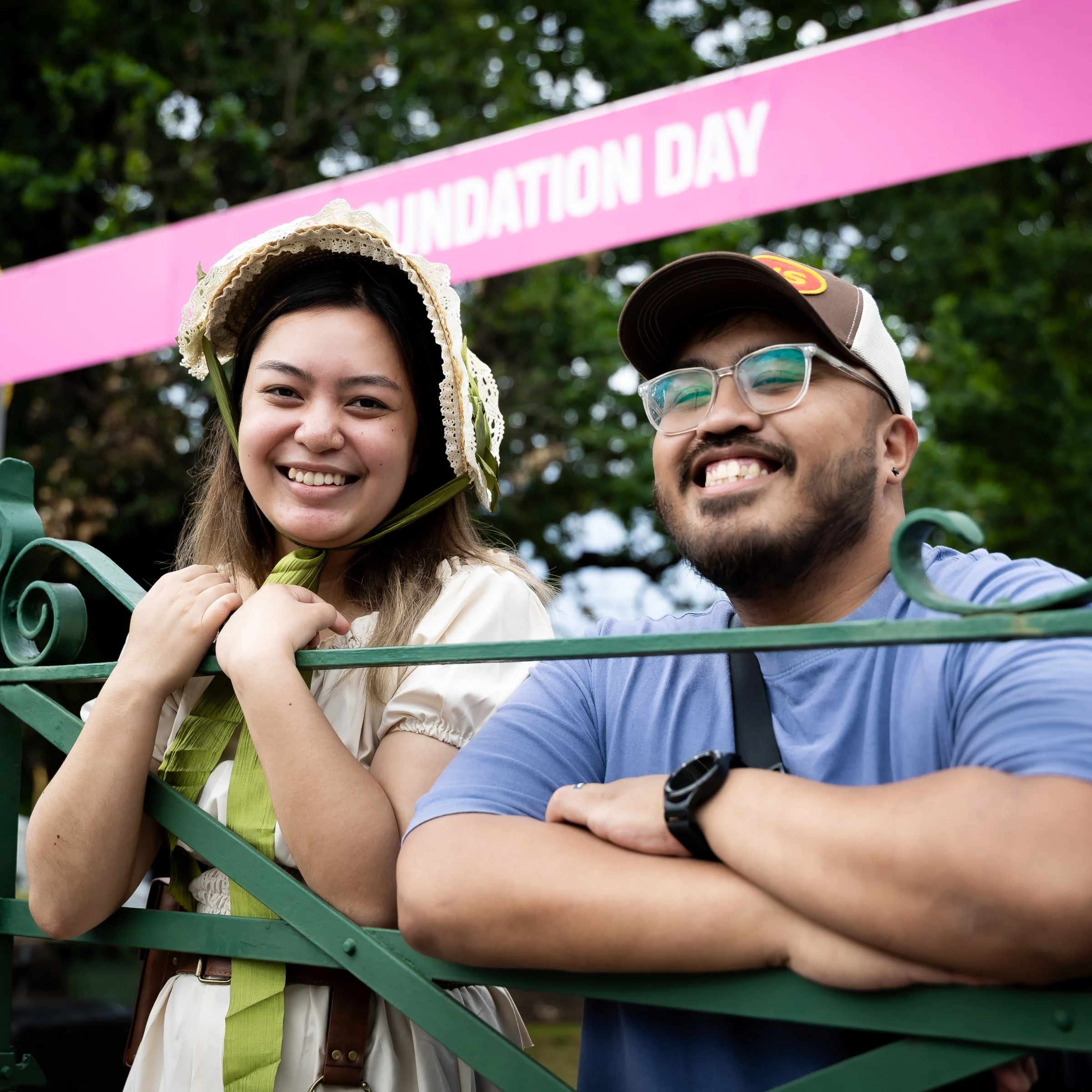 A smiling woman and man pose behind a green metal barrier at an outdoor celebration, with a pink “Foundation Day” banner and leafy trees behind them.