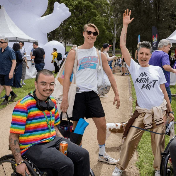 At an outdoor festival, two wheelchair users pose with two standing friends amid colorful tents; one waves beneath a giant white inflatable figure.