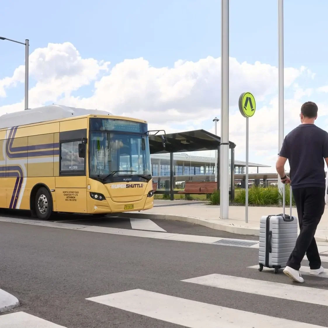 A couple with rolling suitcases crosses at a marked pedestrian crossing as a yellow city bus waits nearby on a sunny day, evoking calm travel.