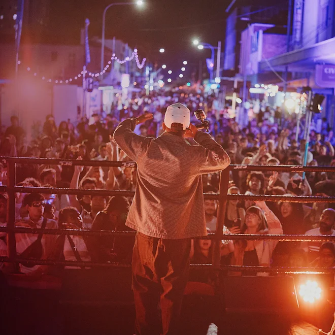A performer in a white cap addresses a packed nighttime street crowd from a stage, raising a hand beneath string lights and colorful spotlights; energetic, festive atmosphere.