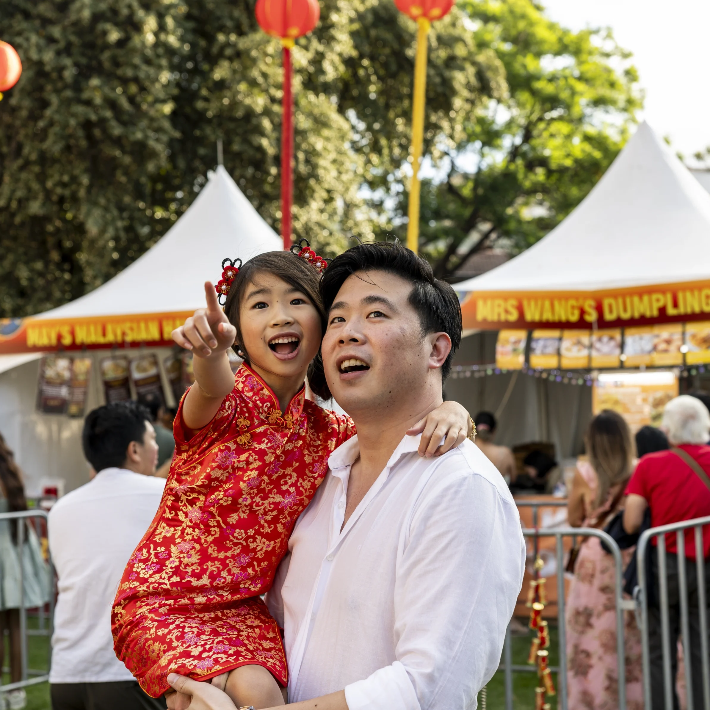 Smiling father carries his daughter in a red Chinese dress as she points at a festival; lanterns and a dumpling stall create a joyful scene.