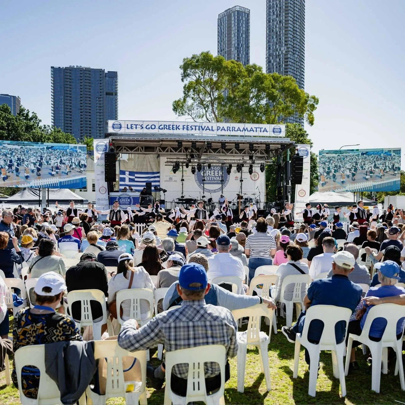 Large outdoor Greek festival in Parramatta, with a cheerful crowd watching dancers on a decorated stage beneath high-rise buildings.