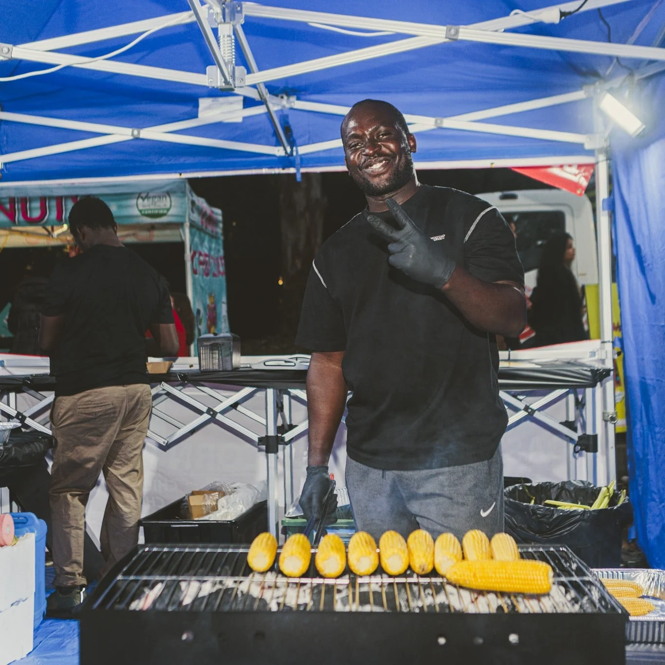 Smiling vendor in black gloves grills rows of golden corn at a lively outdoor food stall beneath a blue canopy, creating a welcoming atmosphere.
