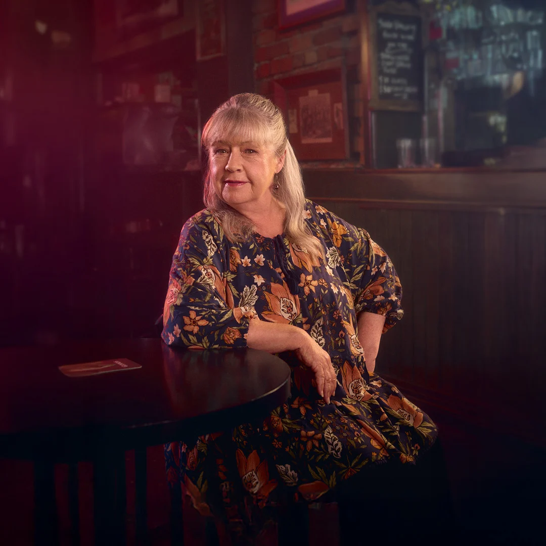 Portrait of a calm, gray-haired woman in a colorful floral dress seated at a pub table, surrounded by dark wood and warm, moody light.