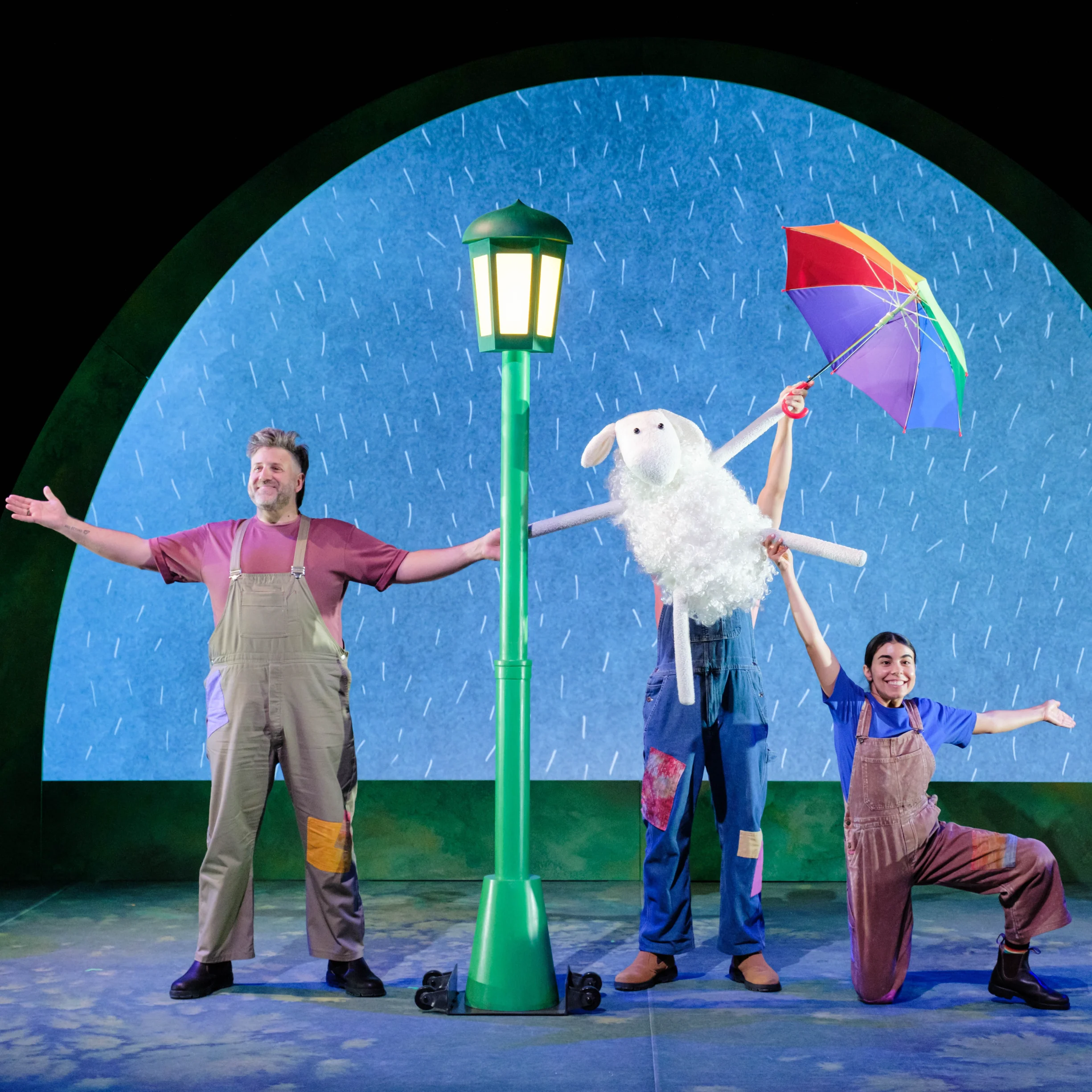 Playful theatrical scene in the rain: three performers surround a green streetlamp, holding a white sheep puppet and bright umbrella.