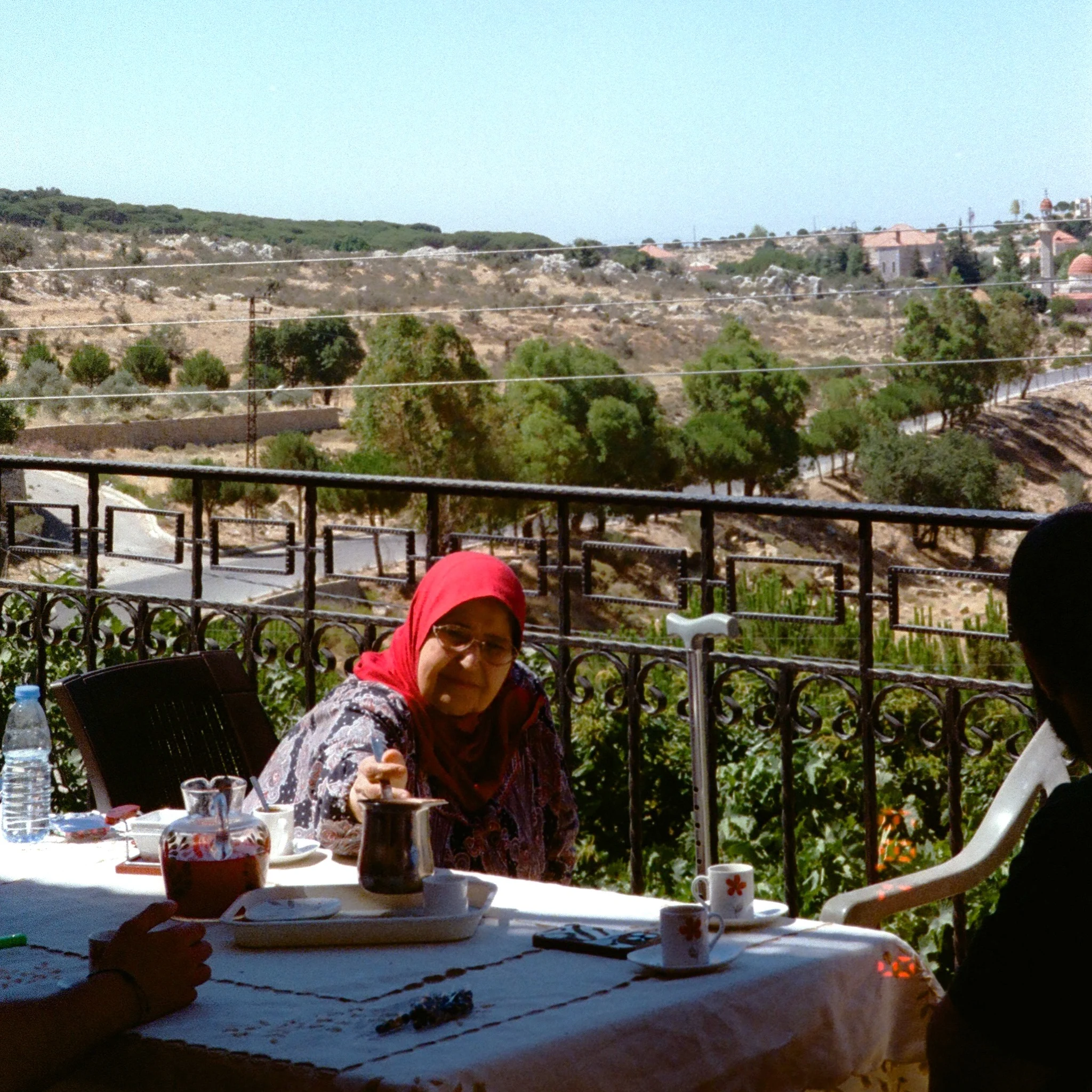 Woman in a red headscarf serves tea at an outdoor table overlooking a sunlit, hilly landscape, creating a warm, peaceful atmosphere.