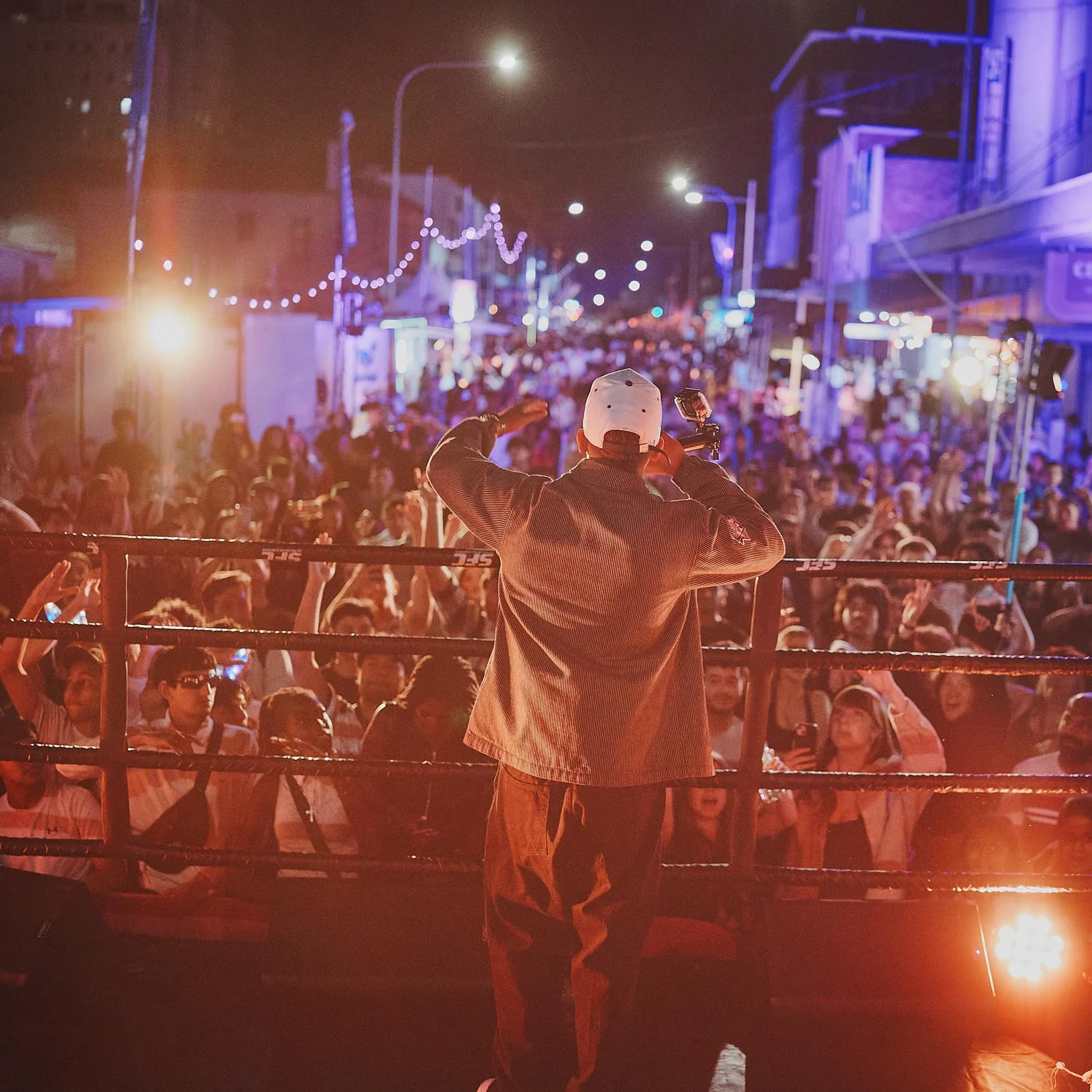 Nighttime street concert: a performer in a white cap faces a cheering crowd, while DJs perform beside him amid colorful city lights.