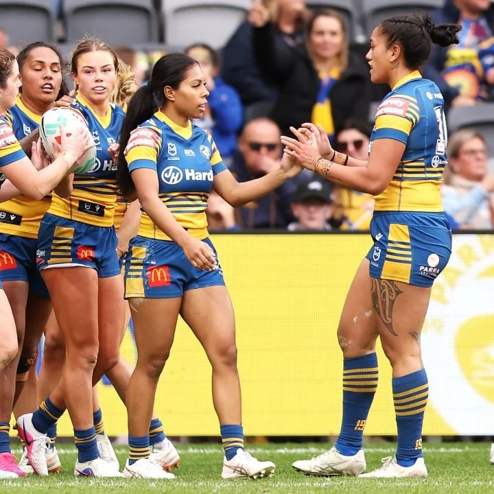 Women’s rugby players in blue-and-yellow uniforms huddle and celebrate on the field, while teammates clap and fans watch behind them.