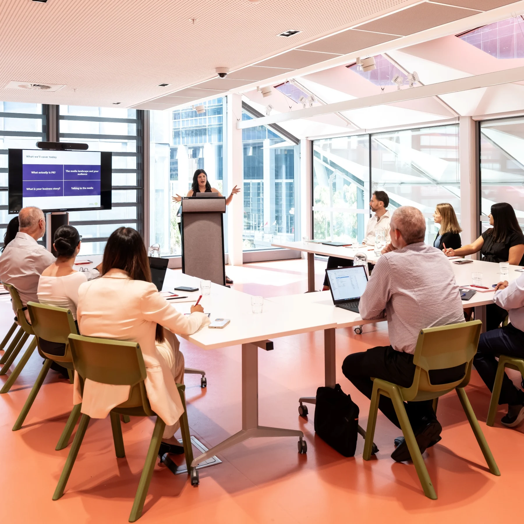 A woman is presenting at a conference table in a bright, modern meeting room with floor-to-ceiling windows. Attendees are engaged, taking notes.