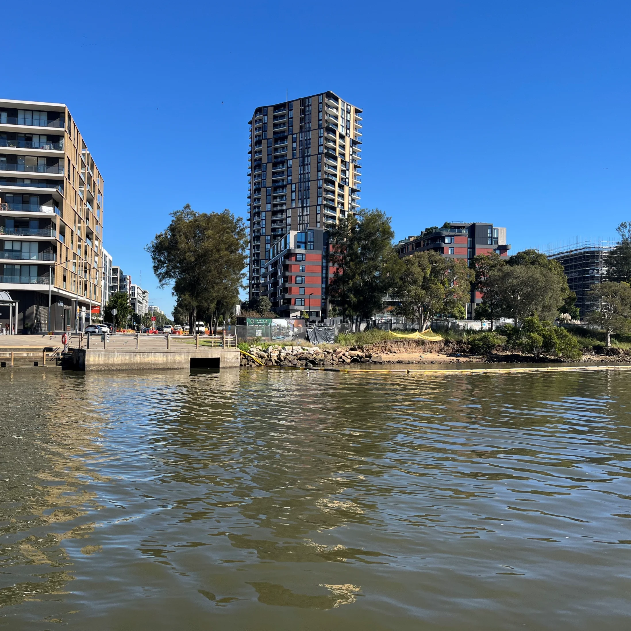 Modern riverside cityscape with high-rise buildings under a clear blue sky. Trees line the riverbank, reflecting in calm water. Relaxed urban setting.