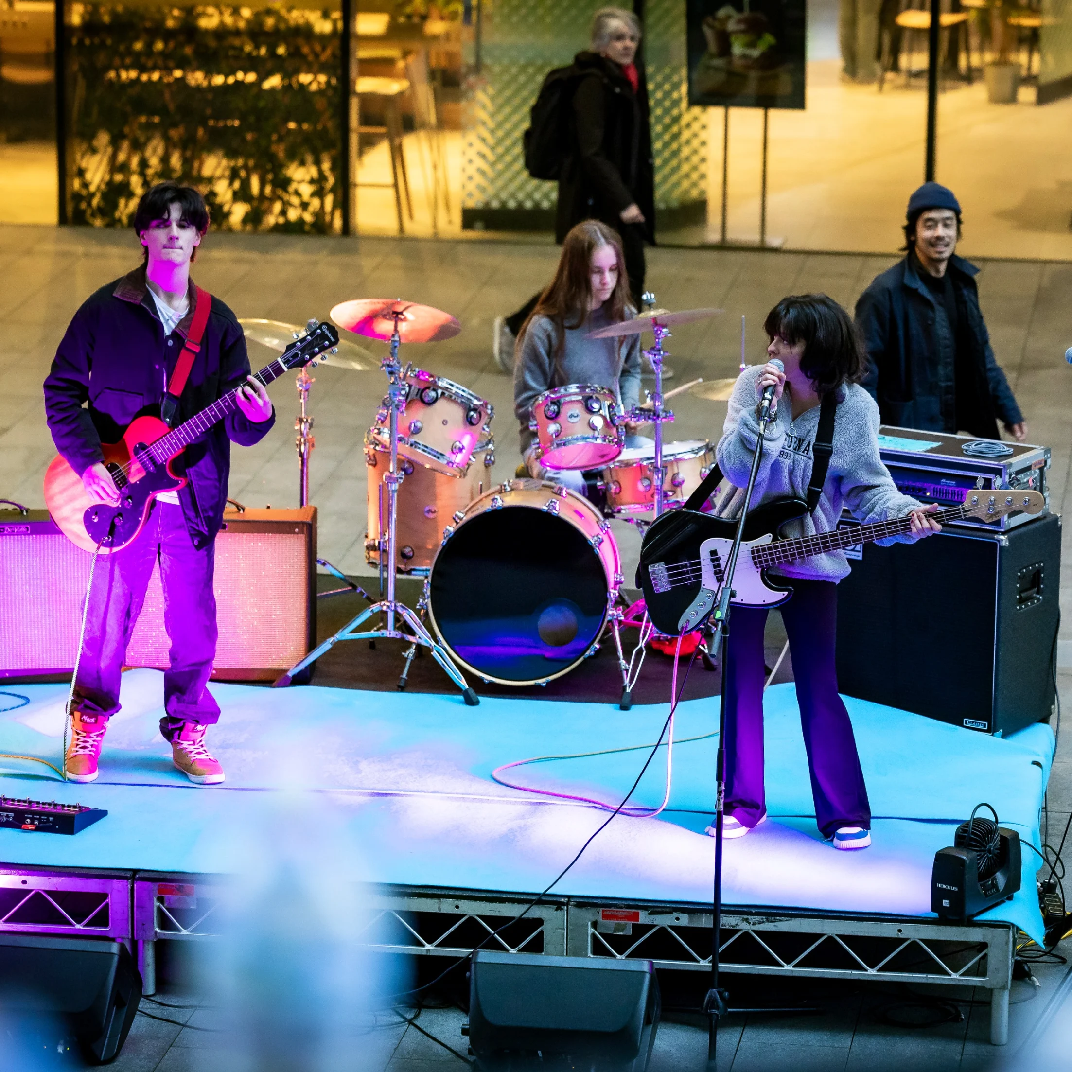 Four adults participate in a yoga class, standinA vibrant indie band performs energetically on an outdoor stage. The guitarist, drummer, and bassist-vocalist play under colorful lights as bystanders watch.g on mats with arms raised, inside a gym with wooden floors and exercise equipment in the background.