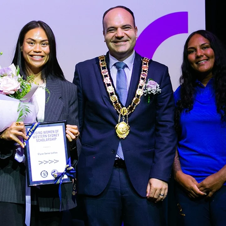 A smiling group of people stands on stage. One holds flowers and a certificate. Another wears a chain of office, suggesting formality and celebration.