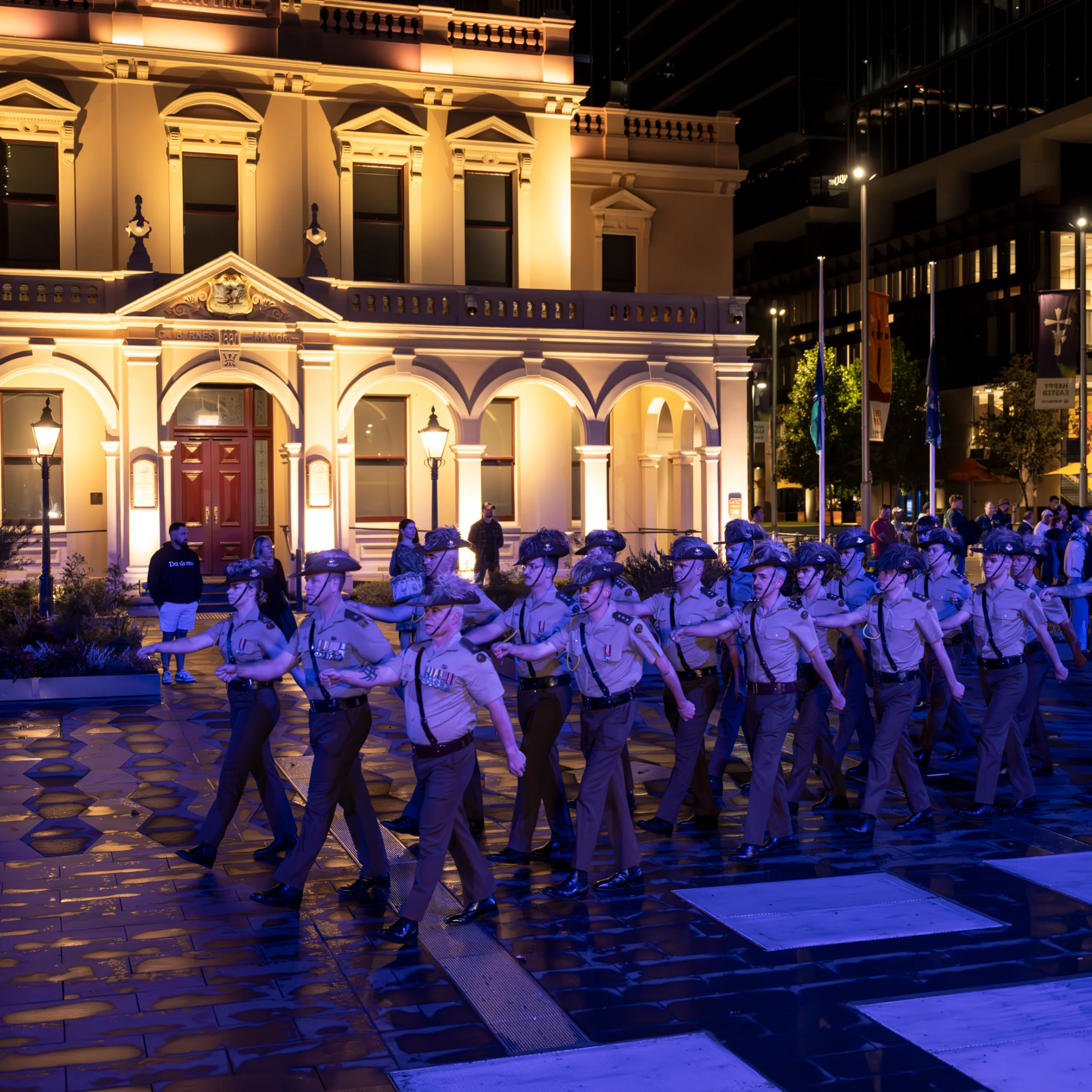 A group of uniformed individuals march in formation on a wet street at night, illuminated by the warm glow of a grand, historic building behind them.