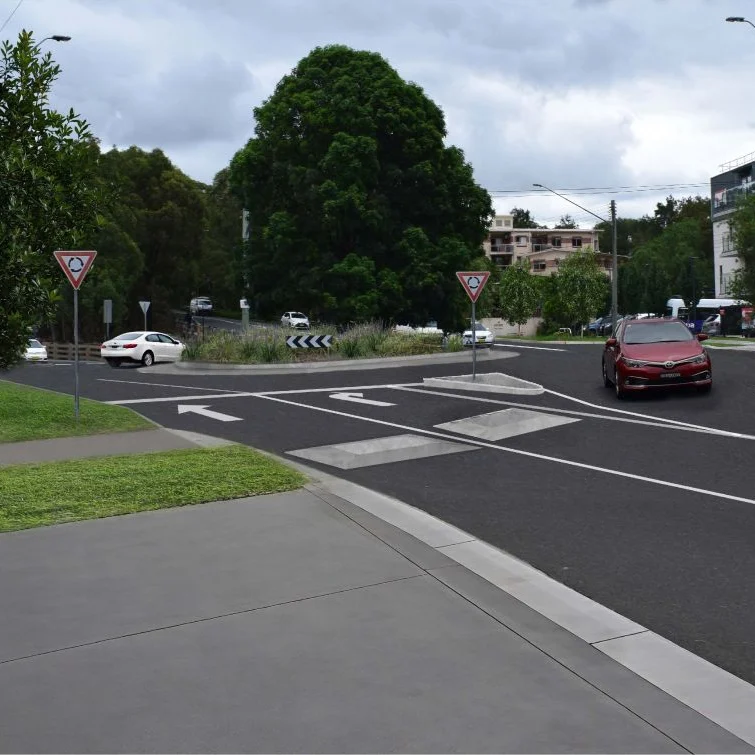 A suburban street scene with a roundabout, trees, and modern buildings. A red car approaches the roundabout, surrounded by green trees under a cloudy sky.