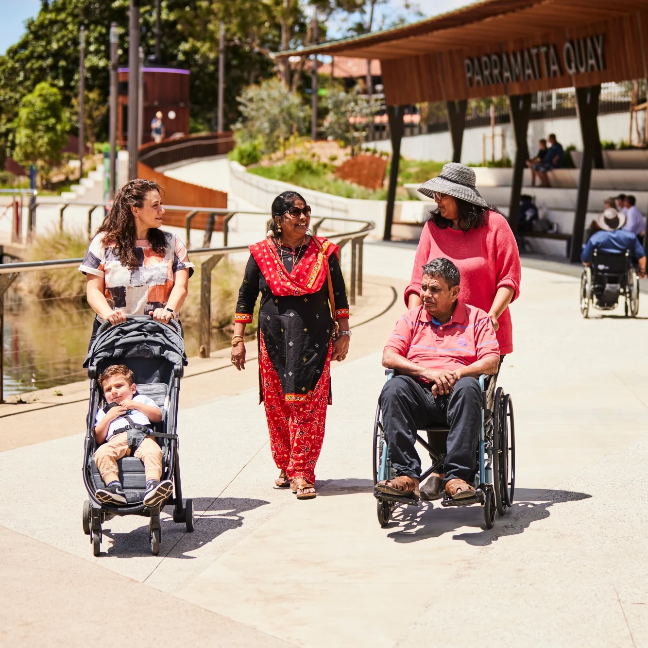 A group enjoys a sunny day at Parramatta Quay. A woman pushes a stroller, while two women talk to a man in a wheelchair, all smiling and engaged.