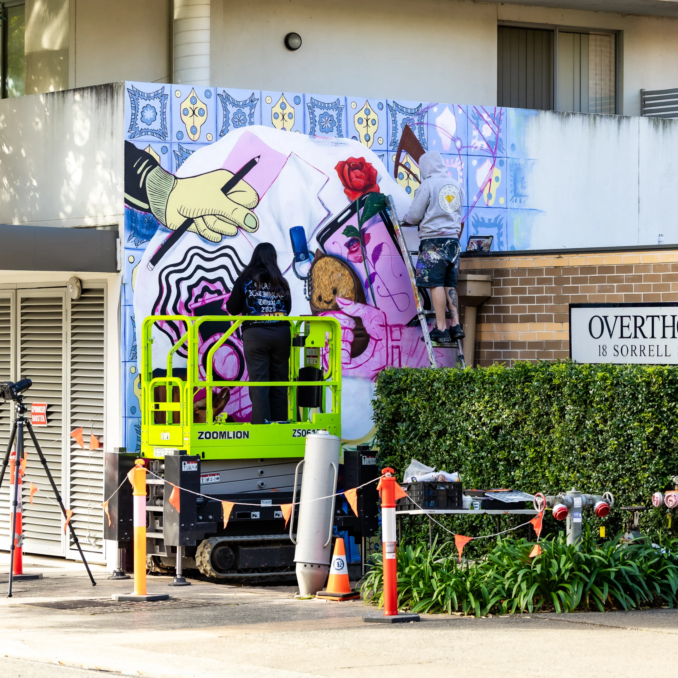 Two individuals on a scissor lift paint a large mural depicting stylized hands, a rose, and abstract shapes on a building exterior, with a street sign reading "OVERTHORPE 18 SORRELL STREET" visible behind a hedge.