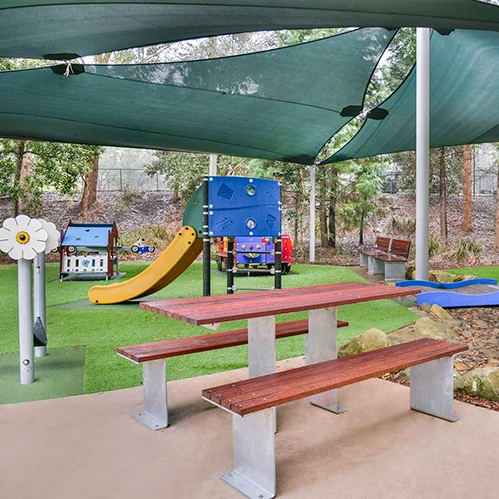 A shaded outdoor playground features a picnic table and benches in the foreground, with various play structures including a slide, climbing wall, and swings in the background.