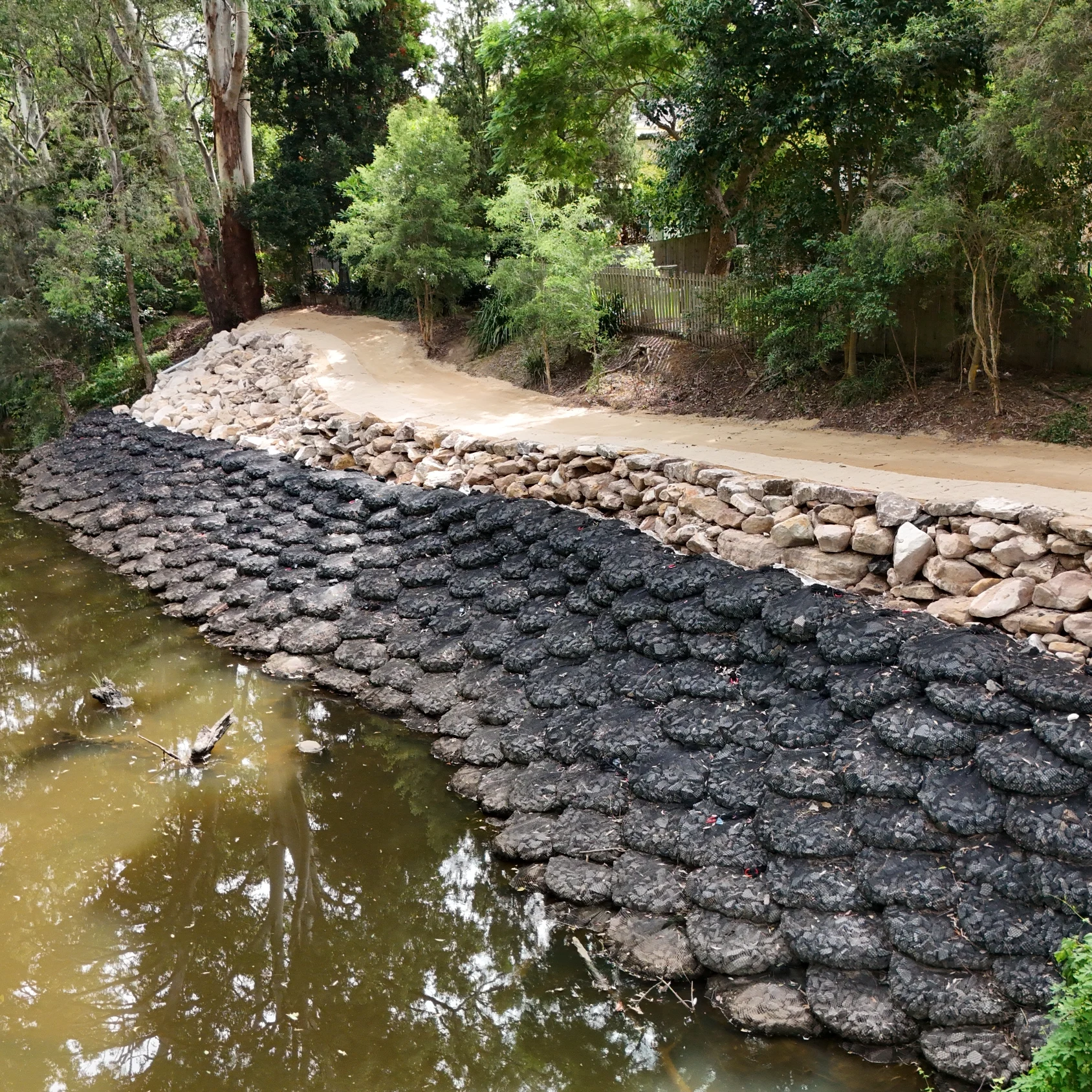 A muddy creek lined with an erosion control barrier of dark, textured interlocking mats, topped with a wall of natural stones, and a sandy path running along the bank.