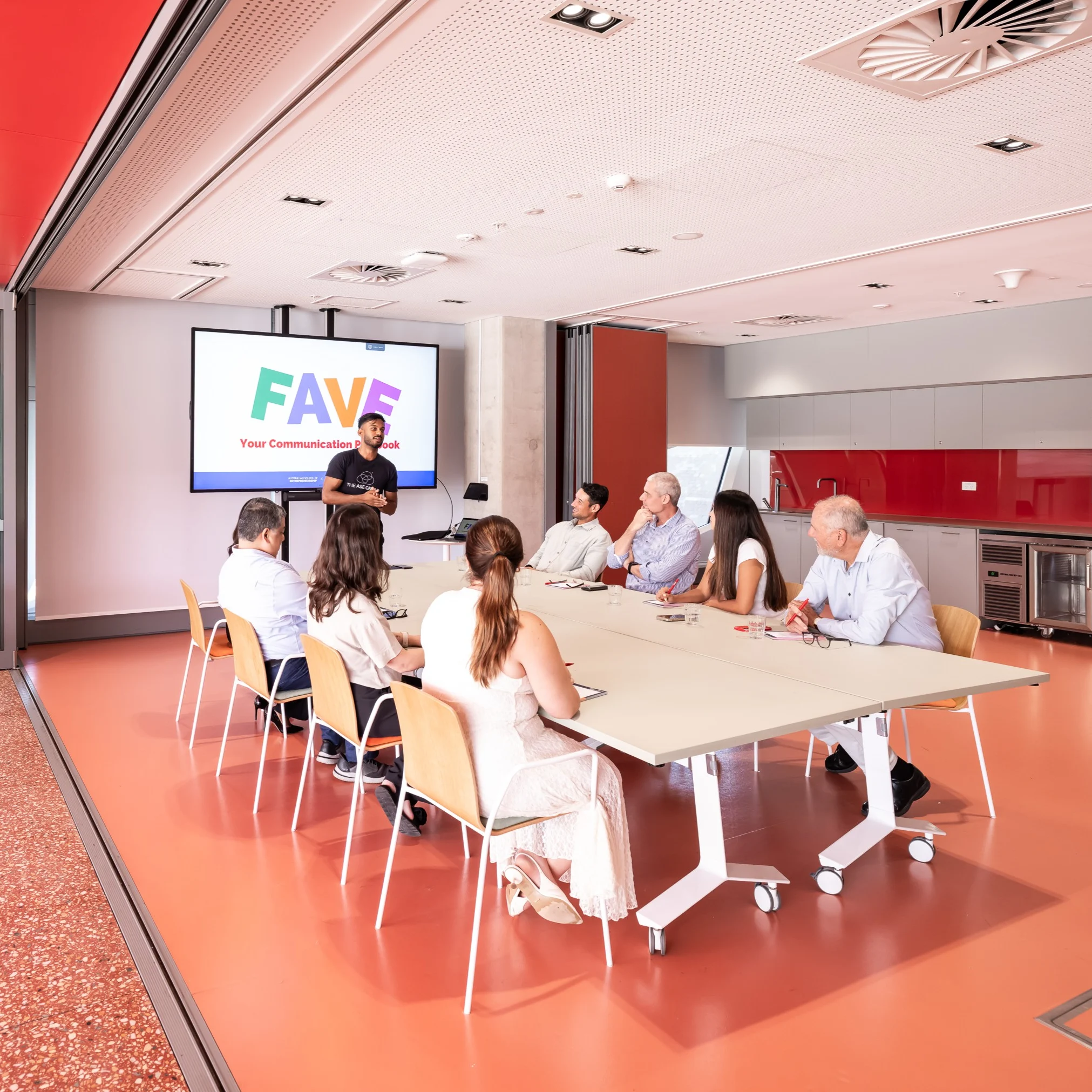 A presenter stands in front of a large screen displaying the word "FAVE" in colorful letters and the text "Your Communication P...Book" below it, addressing a group of people seated around a long conference table.