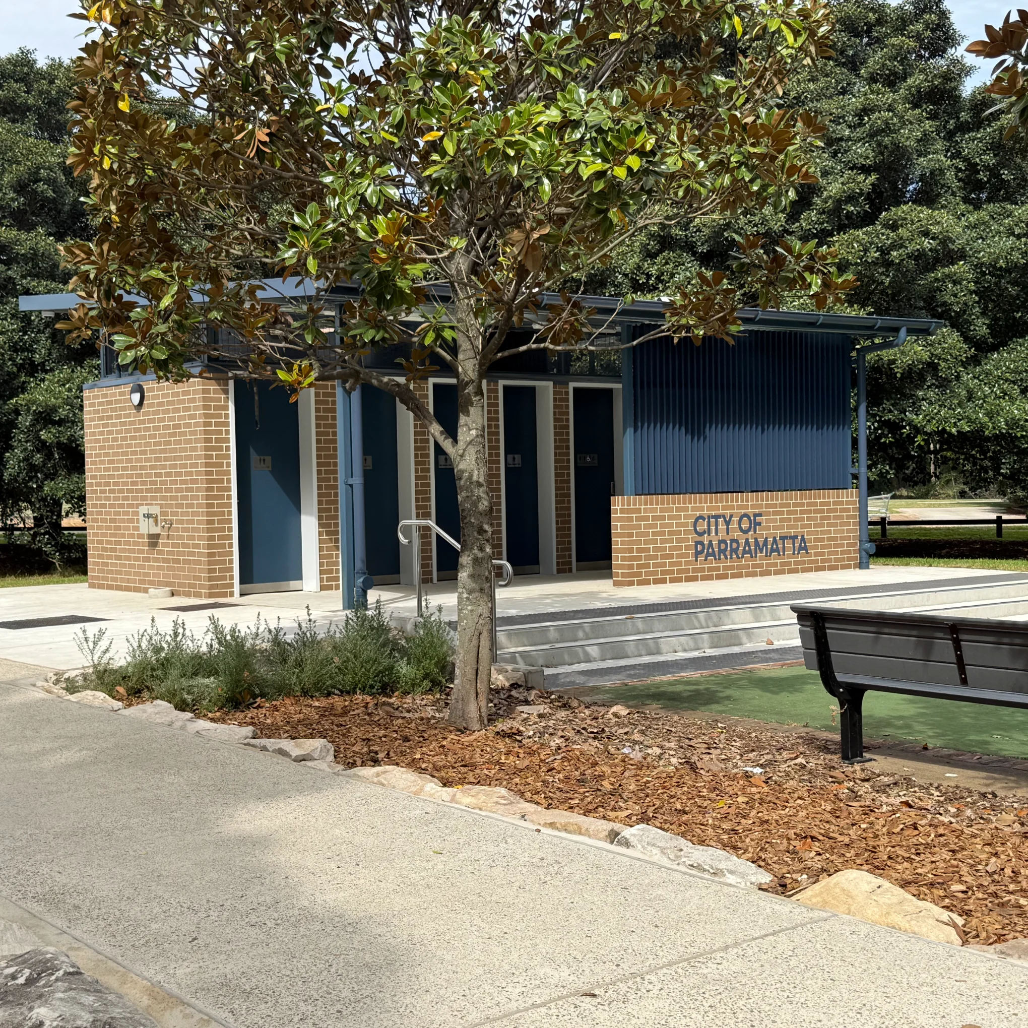 A modern brick and blue public restroom building with "City of Parramatta" signage, situated in a park with mature trees, paved pathways, a bench, and landscaping.