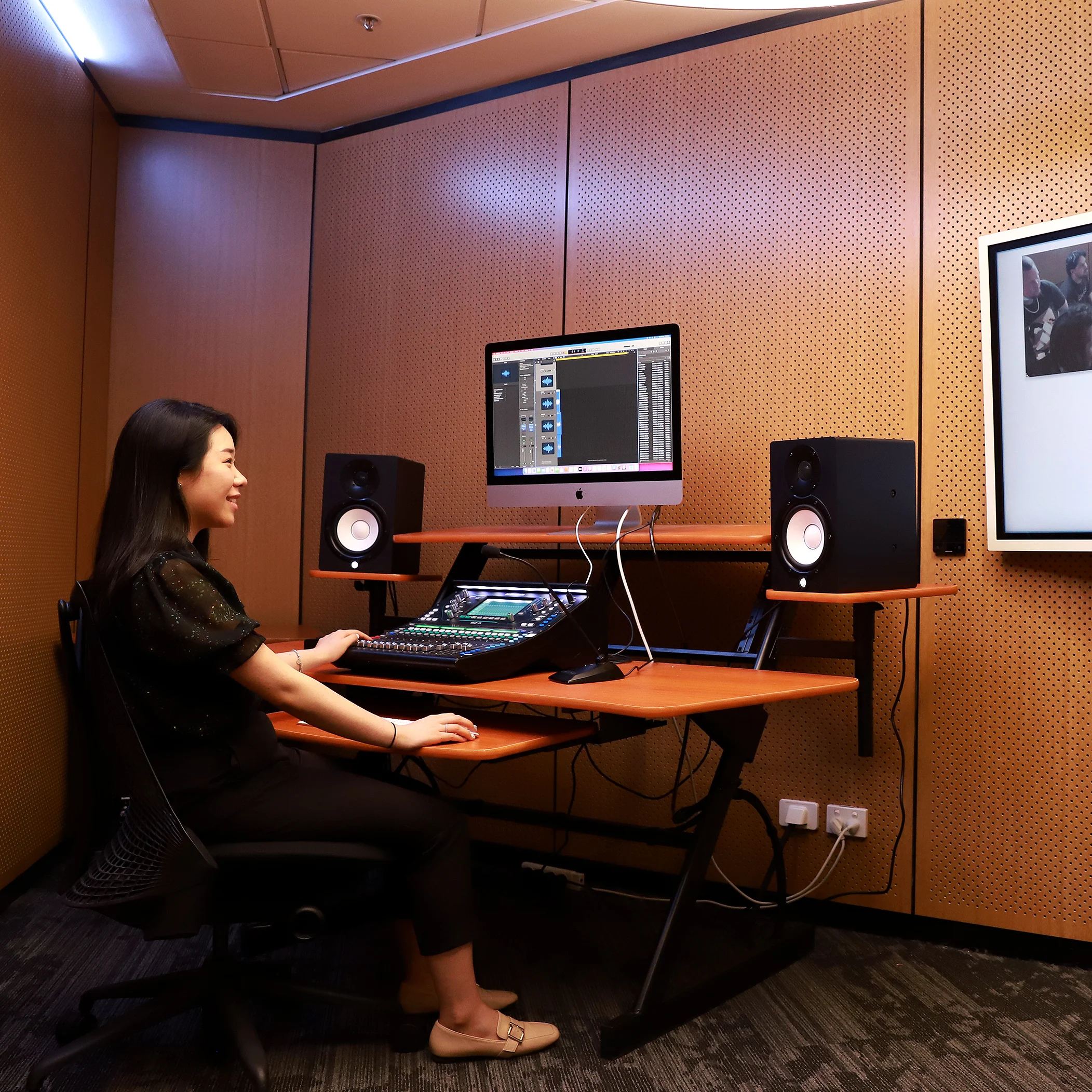 A person works at a mixing board in a sound recording studio, with a computer monitor displaying audio software and a large screen on the wall showing a to-do list and video feeds.