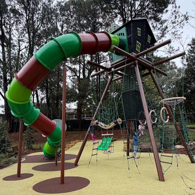 A modern, multi-level playground structure with a large, winding green and brown tube slide, a central climbing tower resembling a black house with green window frames, and various rope nets, ladders, and rings for climbing and play.