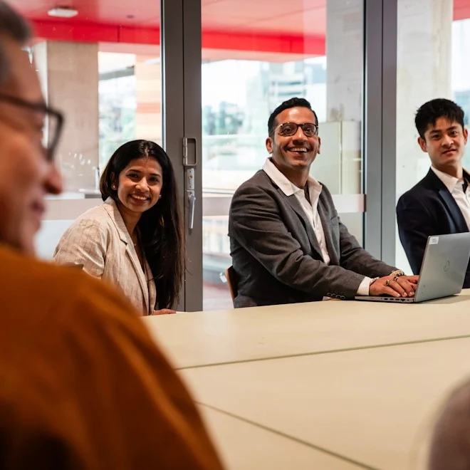 A diverse group of people sits at a table, with one person using a laptop during a meeting in a modern office space.