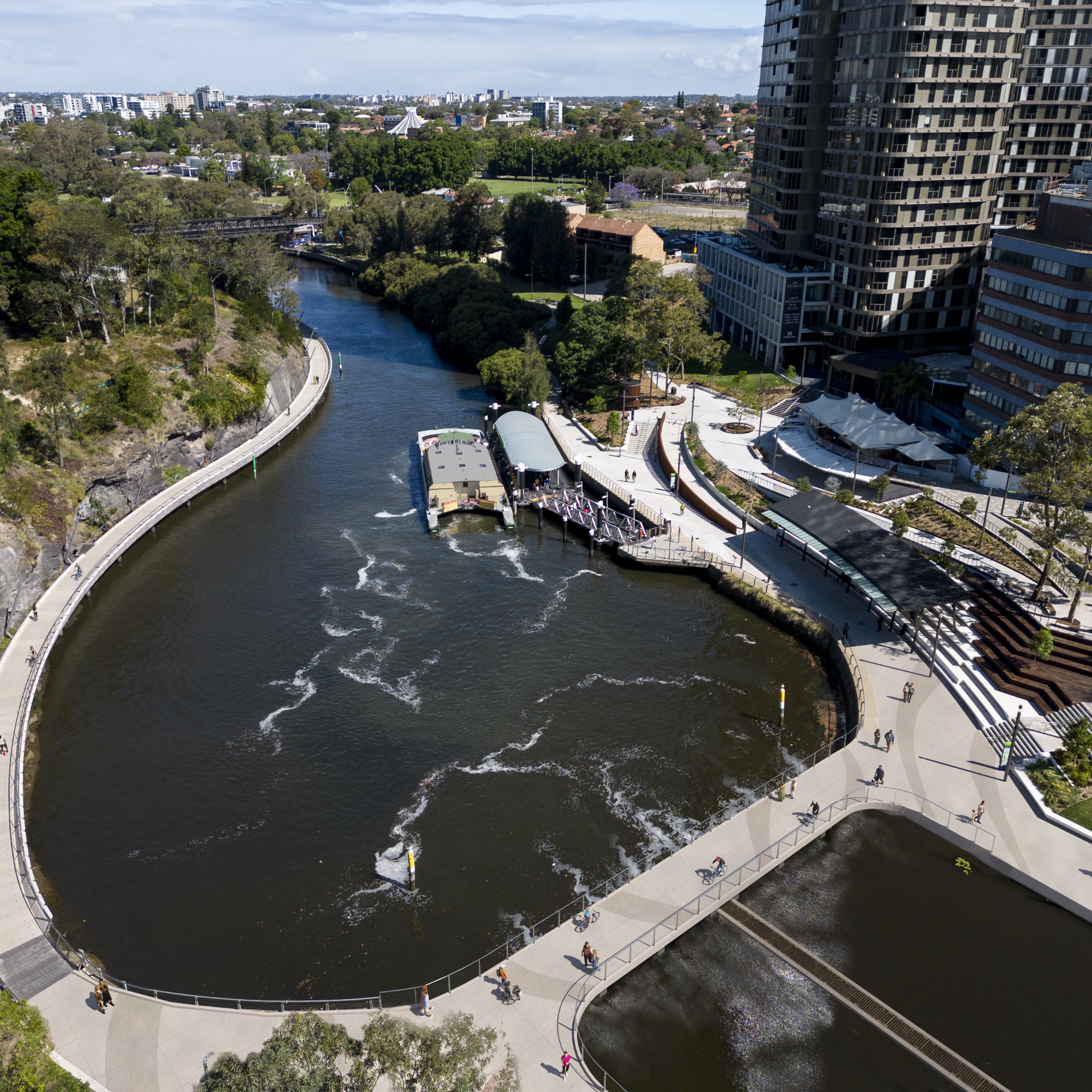 Aerial view of a riverbend surrounded by lush greenery and urban buildings. The river curves through the scene, bordered by walkways with people strolling. The mood is vibrant and dynamic, highlighting urban development and nature's coexistence.