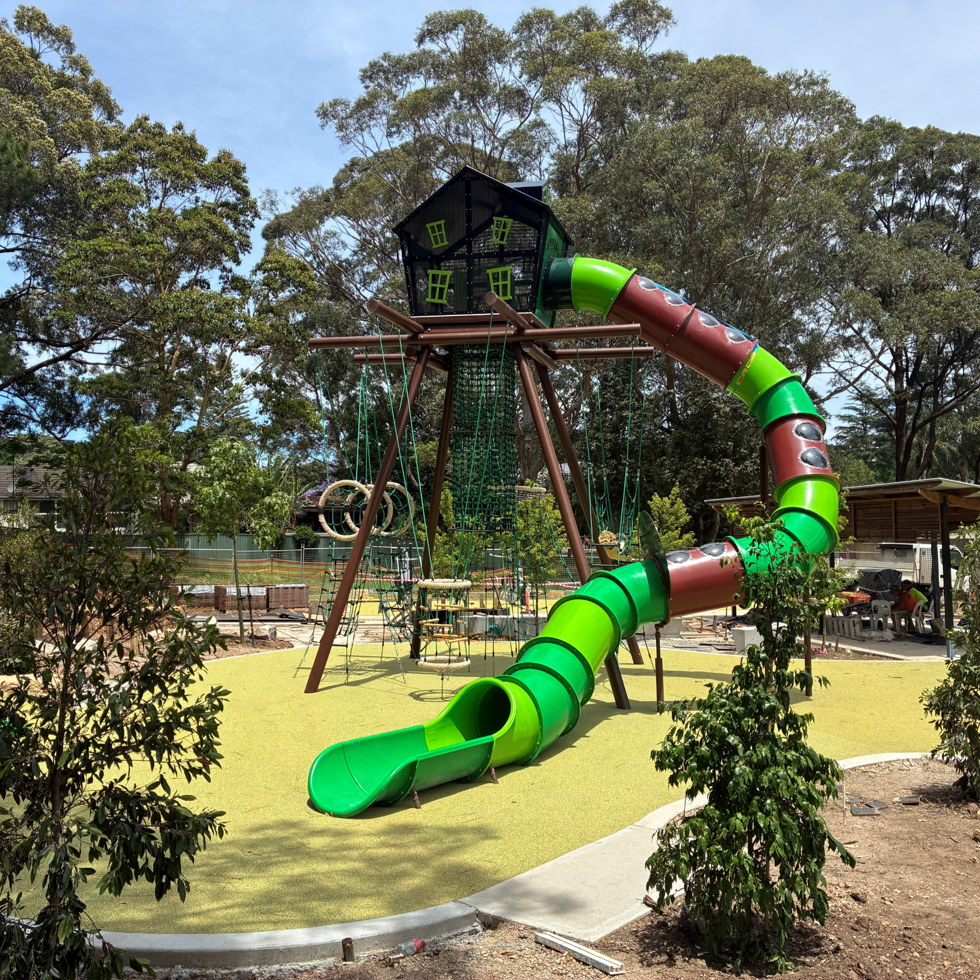 Playground scene with a treehouse and vibrant green, spiral slide, surrounded by trees. The atmosphere is lively and adventurous, inviting for children.