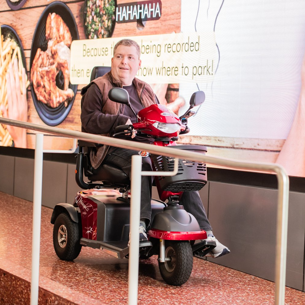 A man in a mobility scooter smiles while ascending a ramp, passing a large screen displaying food images and text, creating a cheerful atmosphere.