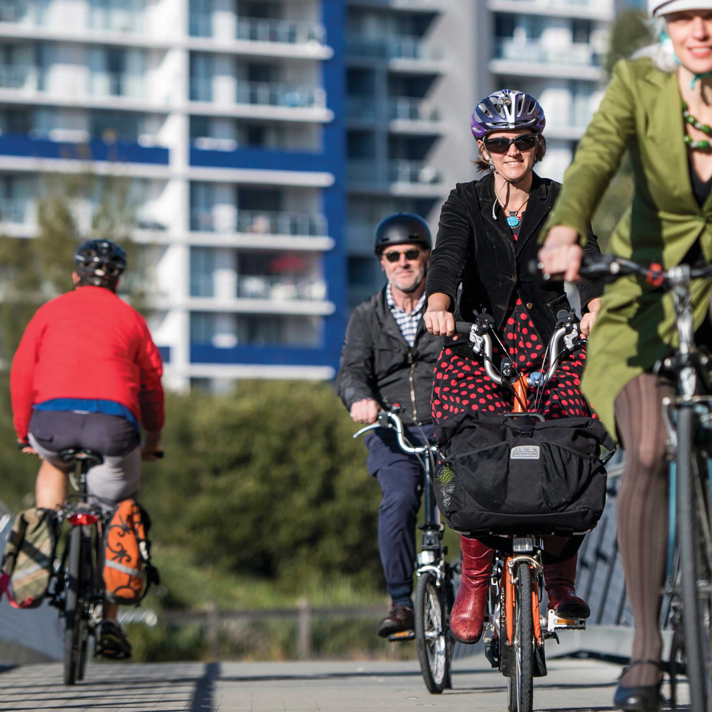 Four people biking on a sunny path, with modern apartment buildings in the background. The scene conveys a lively, urban, and active atmosphere.