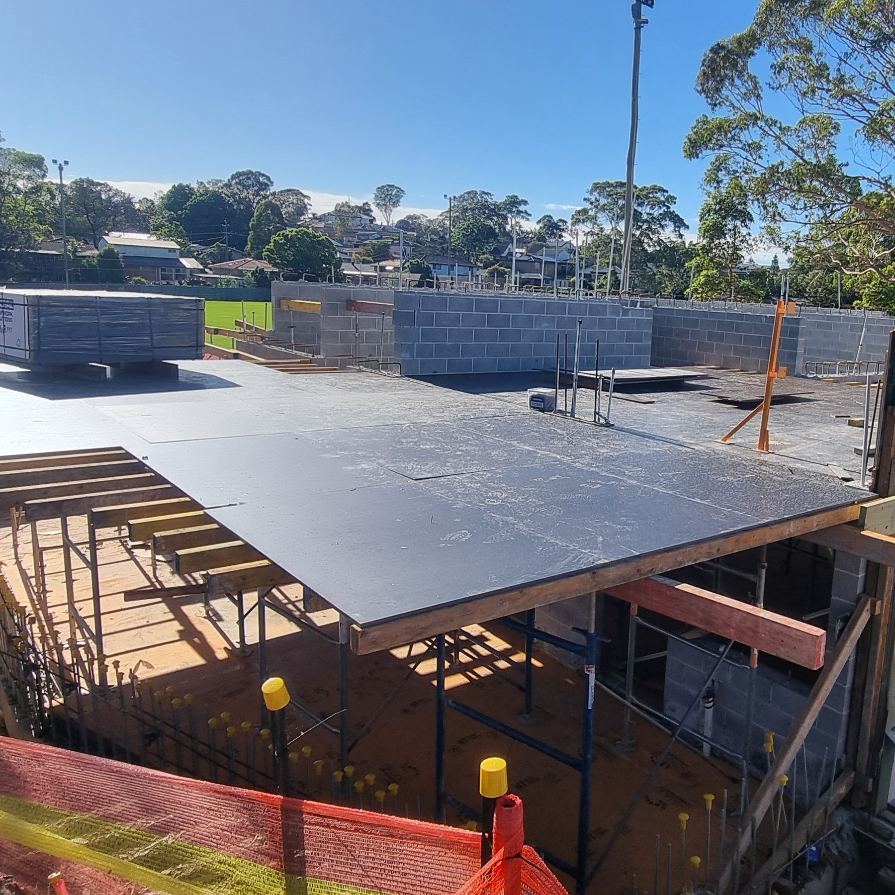 Construction site with unfinished concrete structure surrounded by trees. Sunlit sky in the background and orange safety netting in the foreground.