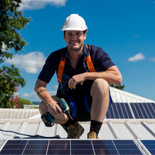 A man in a white hard hat and safety harness squats on a rooftop beside solar panels, under a clear blue sky, holding a tool, smiling confidently.