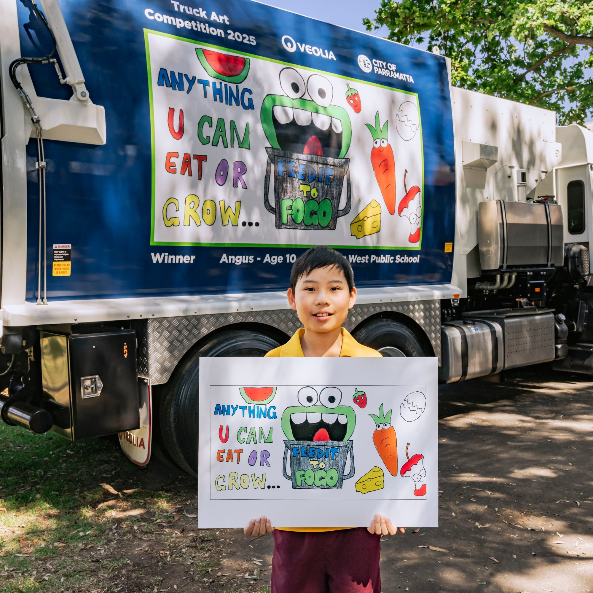 A child proudly holds a colorful poster of a monster with food and text saying "Anything you can eat or grow" in front of a matching garbage truck featuring the same design. The scene is bright and cheerful, conveying excitement and creativity.