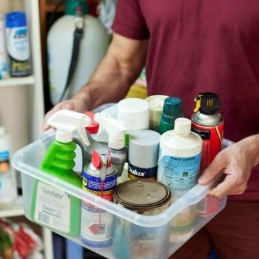 A person in a red shirt holds a plastic bin filled with various cleaning products and aerosol cans. Shelves with more containers are visible in the background.