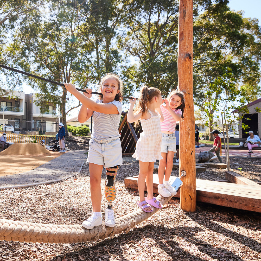 Three smiling girls play on a balancing rope in a sunny playground. One girl uses a prosthetic leg. Trees and play equipment are in the background.