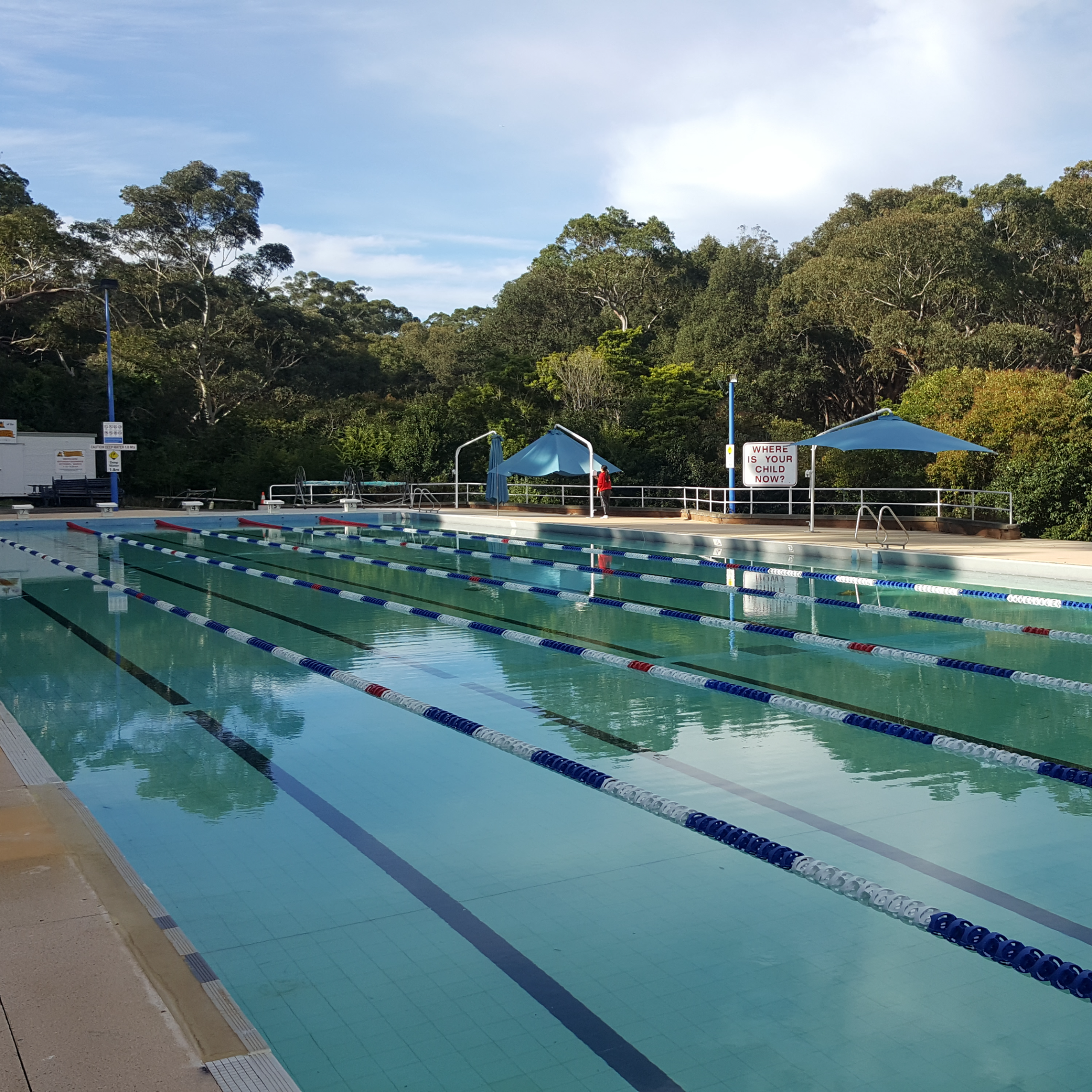 Outdoor swimming pool with clear lanes, surrounded by lush trees and two blue tents. A serene atmosphere under a partly cloudy sky.