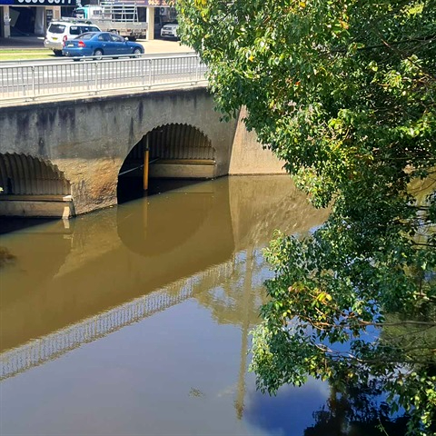 A stone bridge with three arches spans a calm, reflective river. Lush green trees frame the right side, with parked cars visible above the bridge.