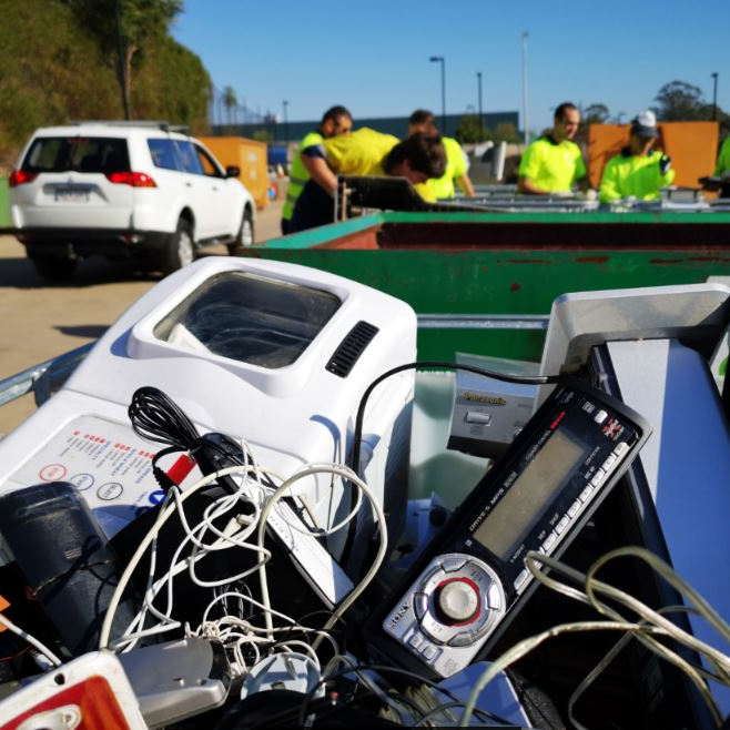 A pile of electronic waste, including gadgets and tangled wires, fills a bin. In the background, people in bright vests sort items near green dumpsters on a sunny day.