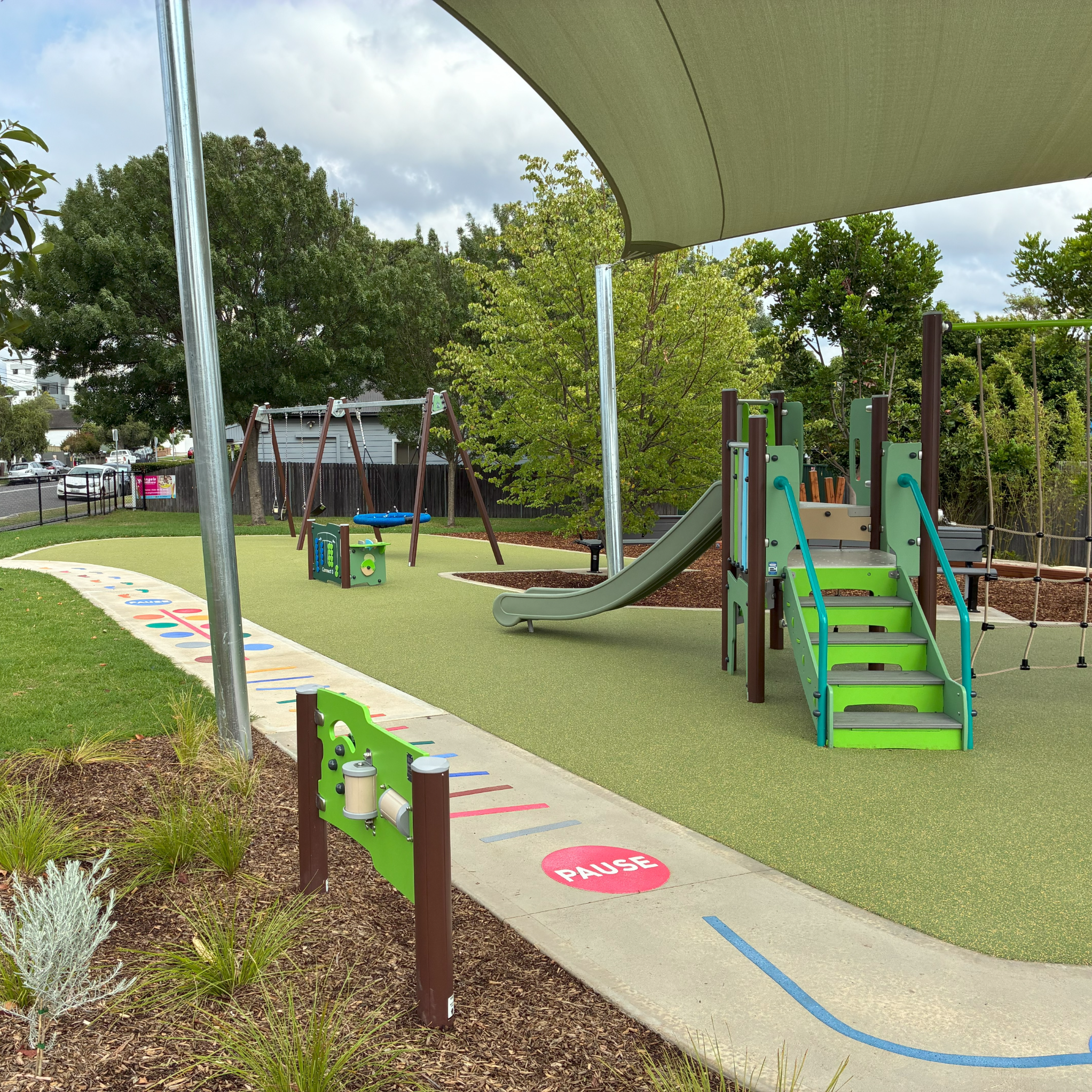 Playground scene with green slides and climbing structure under a canopy. Swings and a small play area are on a soft, green surface. Trees line the park.
