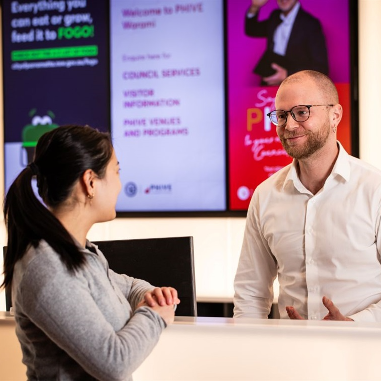 A man in a white shirt talks to a woman in a gray sweater at a reception desk. Digital screens displaying information are visible in the background.