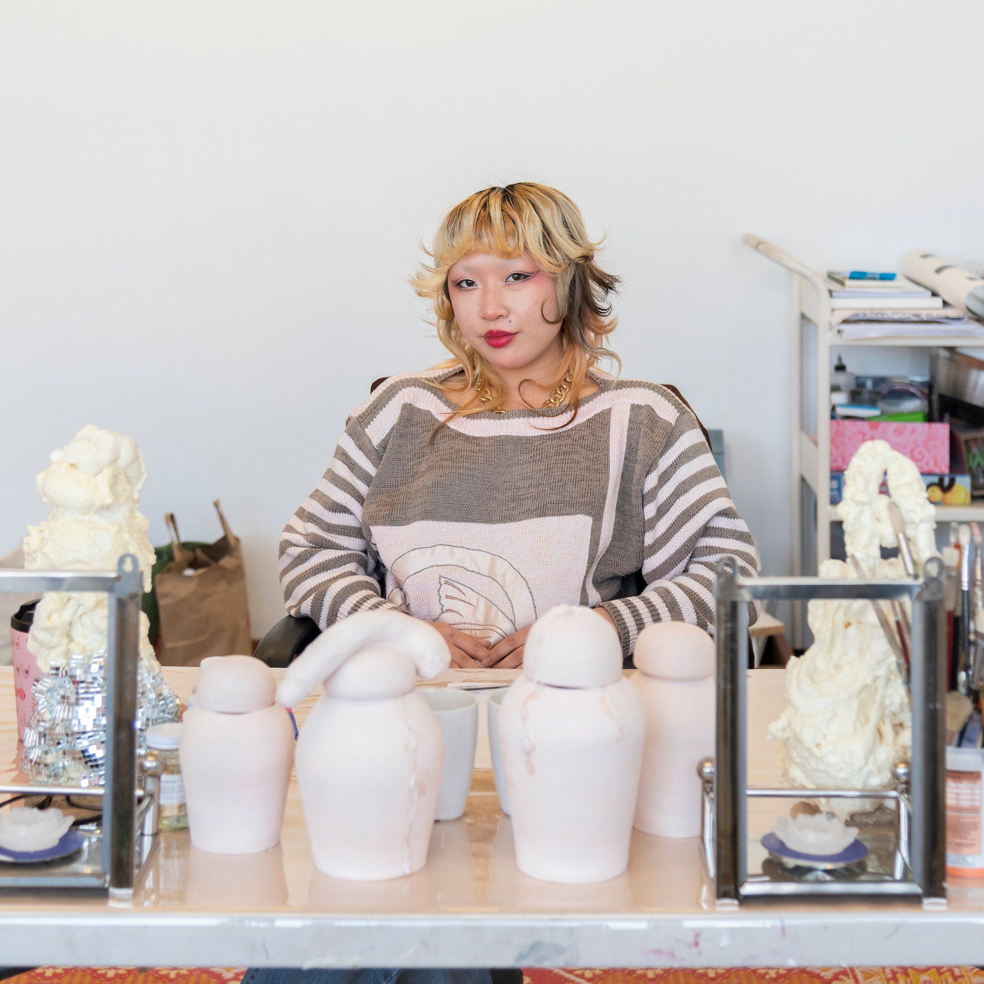 Artist sitting confidently at a cluttered studio table, surrounded by abstract sculptures and art supplies. The mood is creative and focused.