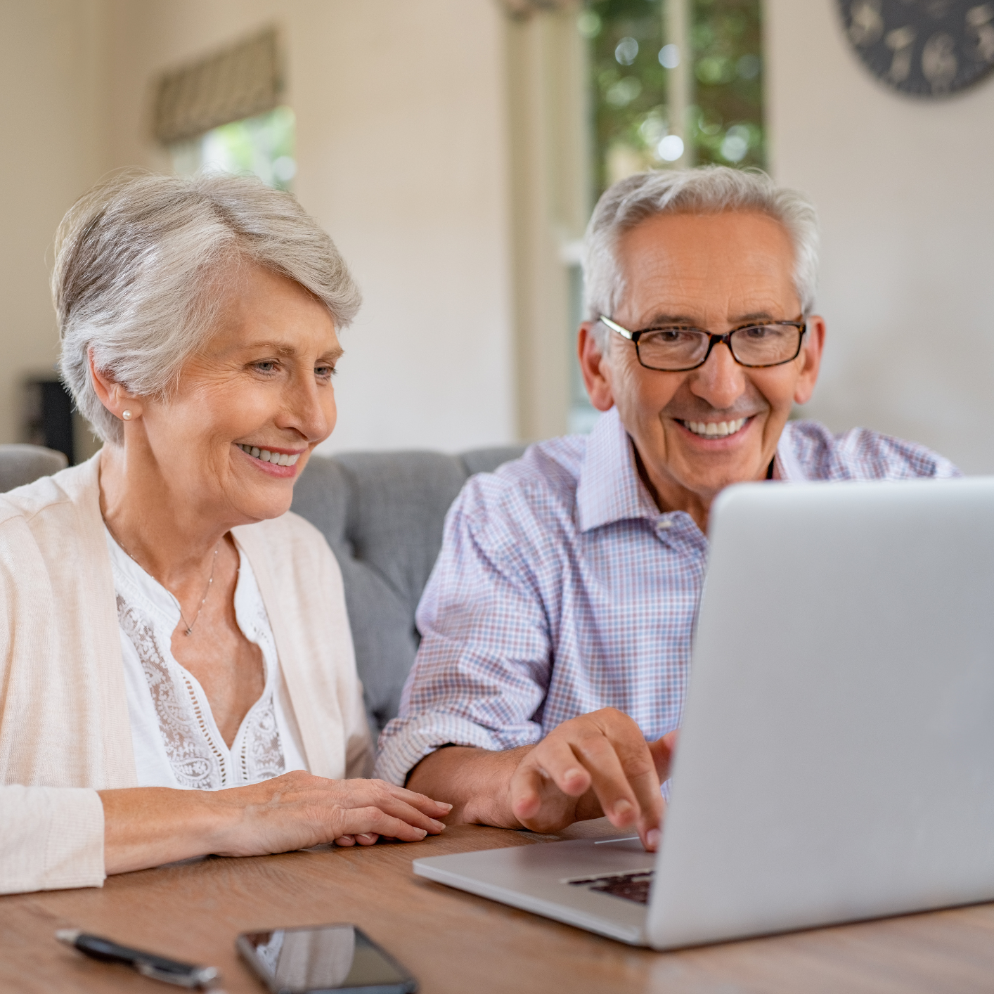Elderly couple smiling while using a laptop at home. The couple appears joyful and engaged, seated on a sofa in a bright, cozy living room.
