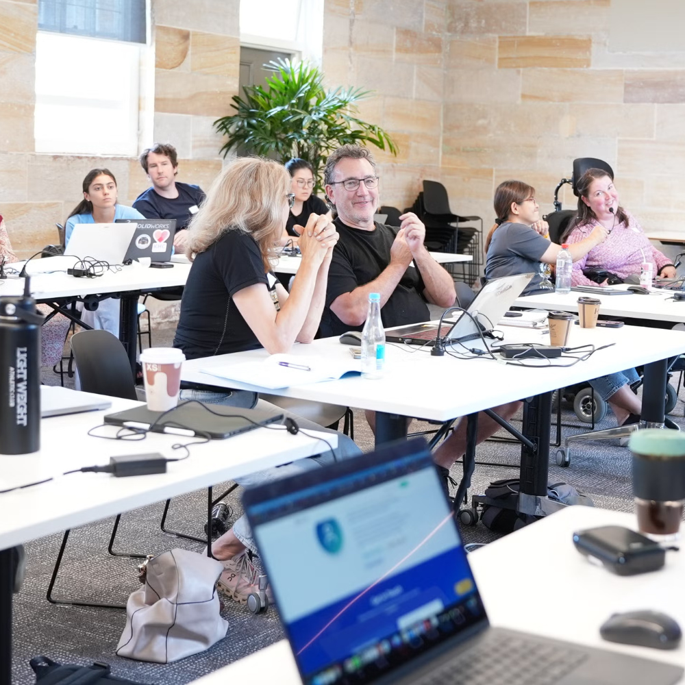 A group of people in a modern classroom with laptops are engaged in discussion. The bright room has large windows and a relaxed, collaborative atmosphere.