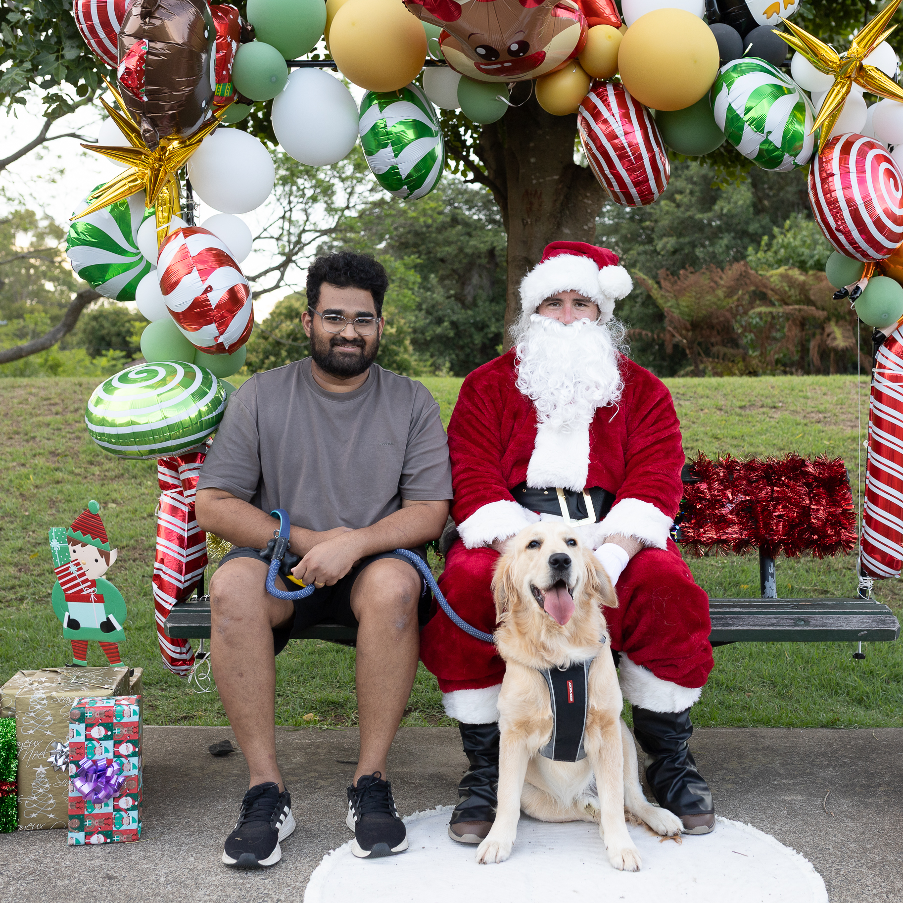 A man with a dog sits next to Santa Claus on a park bench. They're surrounded by festive decorations, including balloons and wrapped gifts. The setting is cheerful and playful.